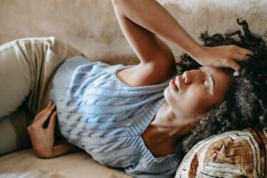 A woman touching her head while lying on her sofa looking to be in pain.