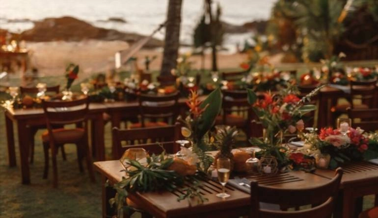 Ξύλινα τραπέζια για δείπνο γάμου διακοσμημένα με τροπικά λουλούδια, ανανάδες, καρύδες και γυάλινα φωτιστικά.