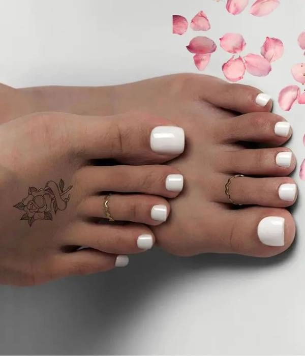 Λευκά νυφικά νύχια ποδιών σε τετράγωνο σχήμα, διακοσμημένα με δύο χρυσά δαχτυλίδια και ένα τατουάζ με τριαντάφυλλο στον αστράγαλο, με ροζ πέταλα λουλουδιών στο φόντο.