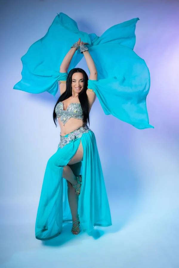 Η Ευδοκία Durrah, καταξιωμένη χορεύτρια Oriental, ποζάρει χαμογελαστή με ένα εκθαμβωτικό μπλε-τιρκουάζ κουστούμι χορού, κρατώντας δύο μεγάλα μπλε πέπλα ψηλά πάνω από το κεφάλι της. Φωτίζεται από ένα απαλό, πολύχρωμο φόντο.