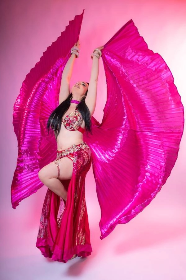 Η Ευδοκία Durrah, καταξιωμένη χορεύτρια Oriental, ποζάρει με μια εντυπωσιακή χορευτική στάση, φορώντας ένα φούξια και χρυσό κουστούμι χορού. Τα χέρια της είναι ψηλά, κρατώντας δύο μεγάλα, πτυχωτά φούξια πέπλα που απλώνονται σαν φτερά, δημιουργώντας ένα εντυπωσιακό σχήμα γύρω της.