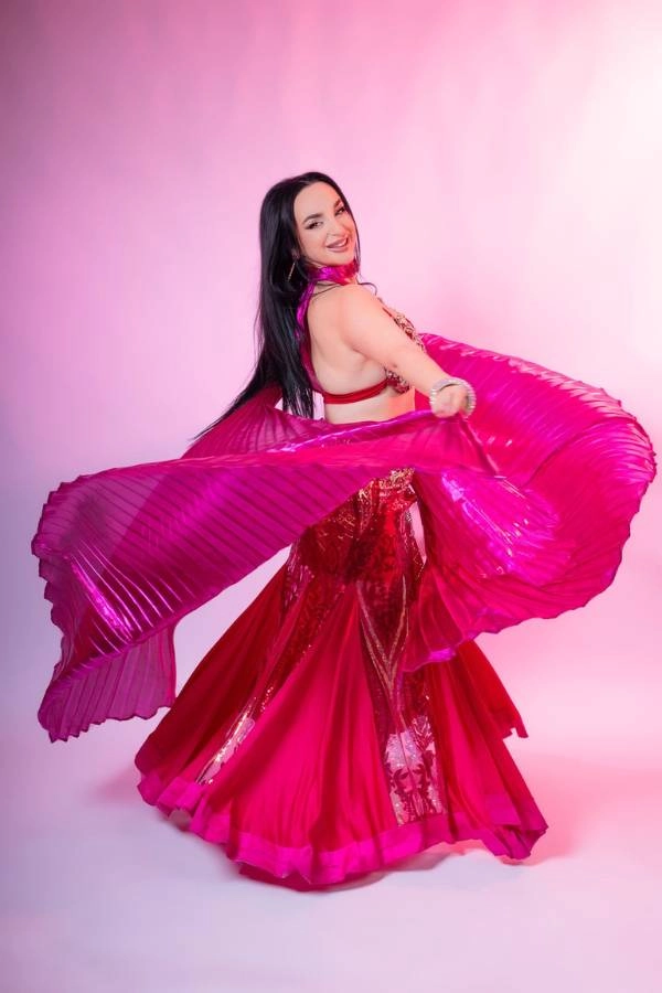 Η Ευδοκία Durrah, καταξιωμένη χορεύτρια Oriental, χαμογελάει και κοιτάζει προς το μέρος του θεατή, φορώντας ένα εντυπωσιακό φούξια κουστούμι χορού.