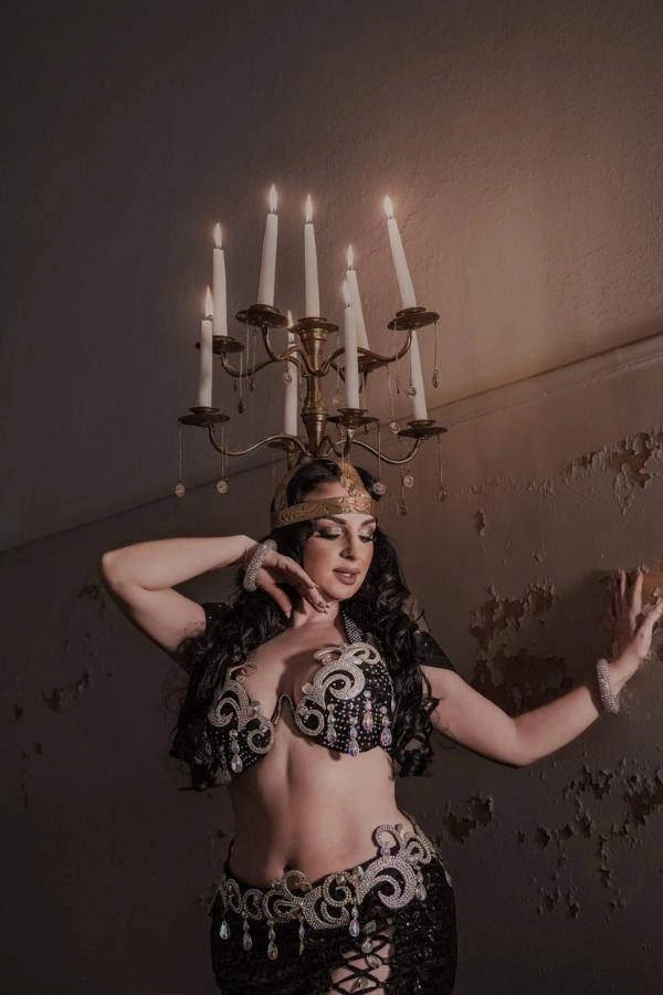 Η Ευδοκία Durrah, χορεύτρια Oriental, ποζάρει σε σκοτεινό, ατμοσφαιρικό φόντο με έναν παλιό τοίχο. Φοράει ένα εντυπωσιακό μαύρο κουστούμι χορού με περίτεχνες ασημί και χρυσές λεπτομέρειες και κεντήματα, ενώ ισορροπεί ένα οκτάφωτο κηροπήγιο με αναμμένα κεριά στο κεφάλι της.