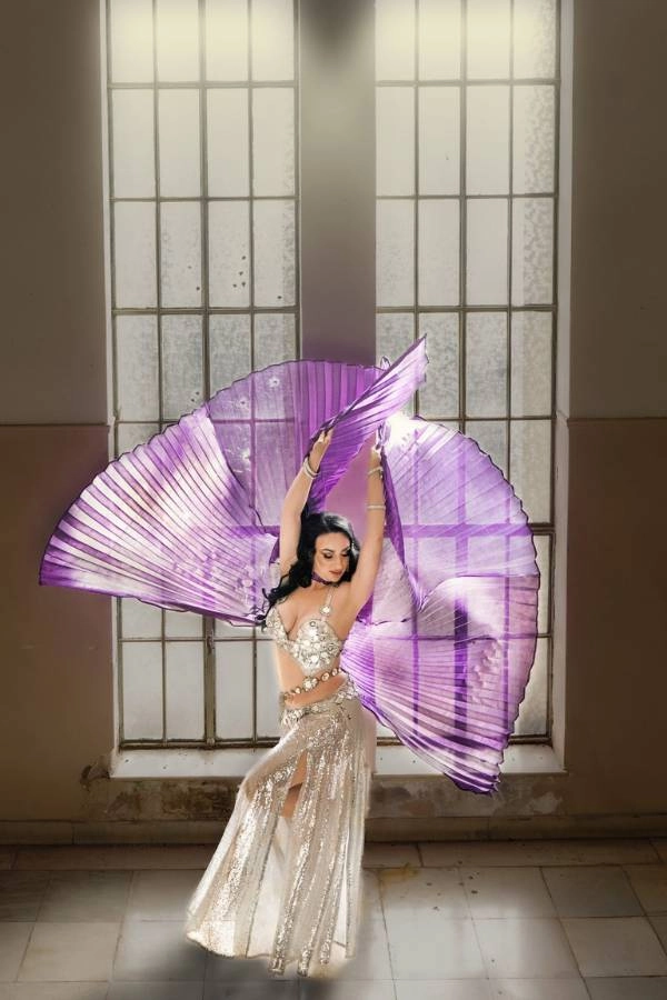 Η Ευδοκία Durrah, καταξιωμένη χορεύτρια Oriental, ποζάρει μπροστά από ένα μεγάλο παράθυρο, φορώντας ένα αστραφτερό, ασημί κουστούμι χορού. Κρατάει ψηλά δύο μεγάλα, πτυχωτά μωβ πέπλα που απλώνονται σαν φτερά πίσω της, δημιουργώντας μια δραματική και εντυπωσιακή εικόνα.