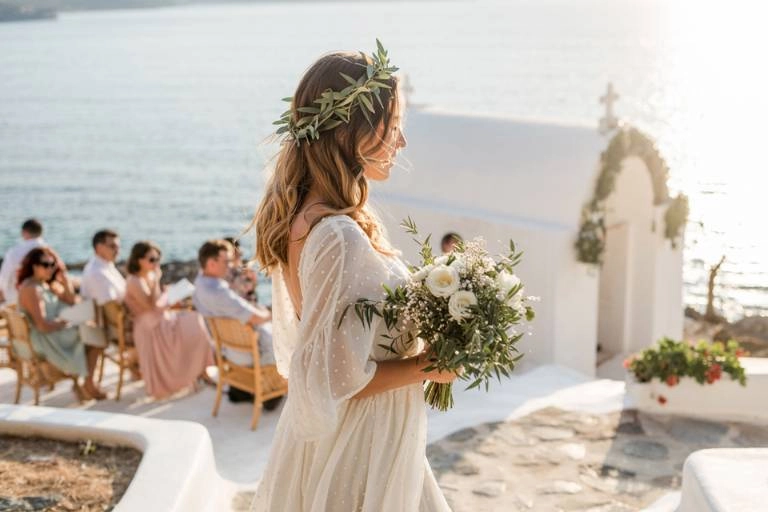Γάμος σε νησί: Οργάνωση, έξοδα, ξωκλήσια, τι να φορέσω! 1 Ονειρεμένος γάμος σε νησί. Νύφη με στεφάνι ελιάς και νυφική ανθοδέσμη περπατά προς ένα λευκό ξωκλήσι δίπλα στη θάλασσα, με καλεσμένους να παρακολουθούν.