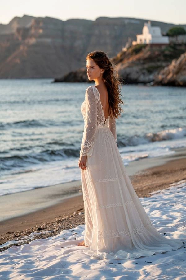 Γάμος σε νησί: Οργάνωση, έξοδα, ξωκλήσια, τι να φορέσω! 8 Μια νύφη με ένα αέρινο νυφικό στέκεται στην αμμουδιά καθώς δύει ο ήλιος, κοιτώντας τη θάλασσα, με ένα γραφικό ξωκλήσι να διακρίνεται στο λόφο στο βάθος, εικόνα που αποτυπώνει την ομορφιά ενός νησιώτικου γάμου.