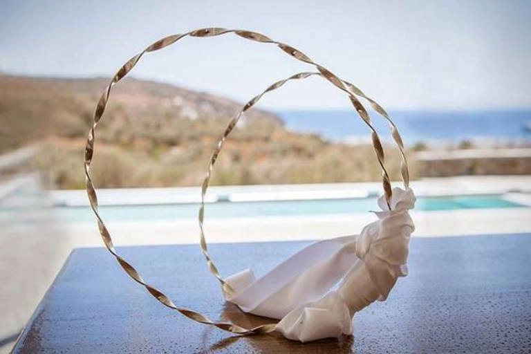 Έξοδα κουμπάρου και υποχρεώσεις | Τι πληρώνει στο γάμο; 4 Στέφανα γάμου τοποθετημένα σε ξύλινη επιφάνεια με φόντο τη θάλασσα και το ελληνικό τοπίο