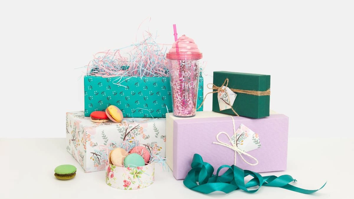 Πολύχρωμα δώρα τυλιγμένα με διακοσμητικά χαρτιά, κορδέλες και κάρτες, συνοδευμένα από macarons και ποτήρι με glitter, σε λευκό φόντο.