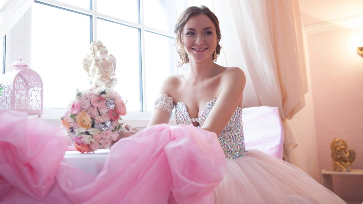 Νύφη με ρομαντικό φόρεμα και ροζ τούλινη φούστα, κάθεται χαμογελαστή δίπλα σε ανθοδέσμη με παστέλ λουλούδια, δίπλα σε παράθυρο.