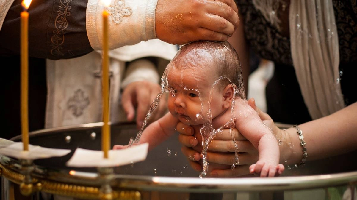 Μωρό την ώρα που βαπτίζεται σε ορθόδοξη τελετή, με ιερέα να ρίχνει νερό στο κεφάλι του και κεριά γύρω από την κολυμπήθρα.