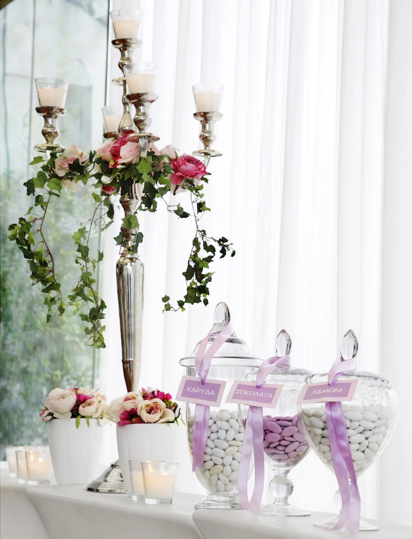 Candy bar γάμου με γυάλες και πινακίδες γεύσεων, στολισμένο με ροζ τριαντάφυλλα, κηροπήγιο και αρωματικά κεριά, από τη Vassiliki Creations