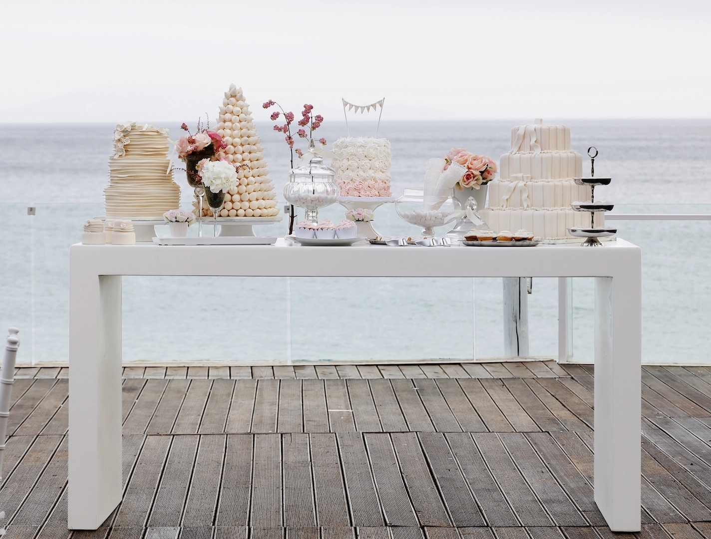 Εντυπωσιακό τραπέζι γλυκών γάμου με θαλασσινό φόντο, τούρτες και ρομαντικά λουλούδια από τη Vassiliki Creations