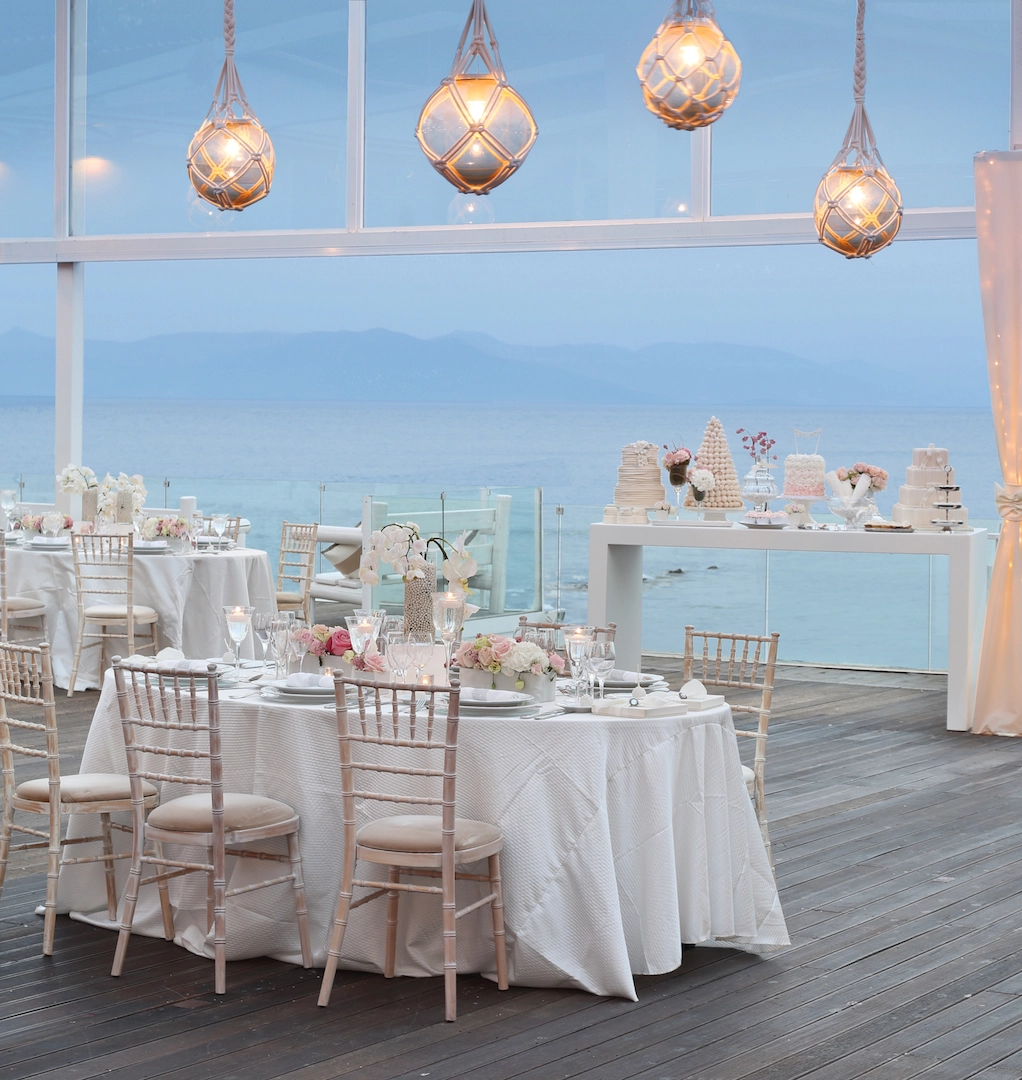 Ρομαντικό σκηνικό δεξίωσης δίπλα στη θάλασσα με λευκά τραπέζια, ροζ λουλούδια και φωτιστικά ρομαντικού στυλ από τη Vassiliki Creations