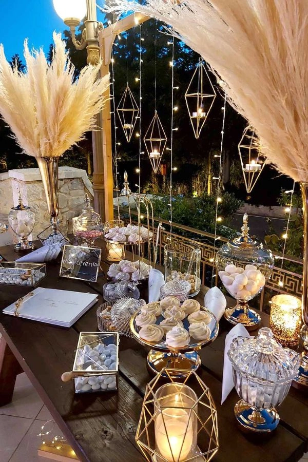 Boho glam στολισμός δεξίωσης με pampas grass, χρυσές λεπτομέρειες, κεράκια και γυάλες σε ξύλινο τραπέζι, δημιουργώντας ζεστή και λαμπερή ατμόσφαιρα.