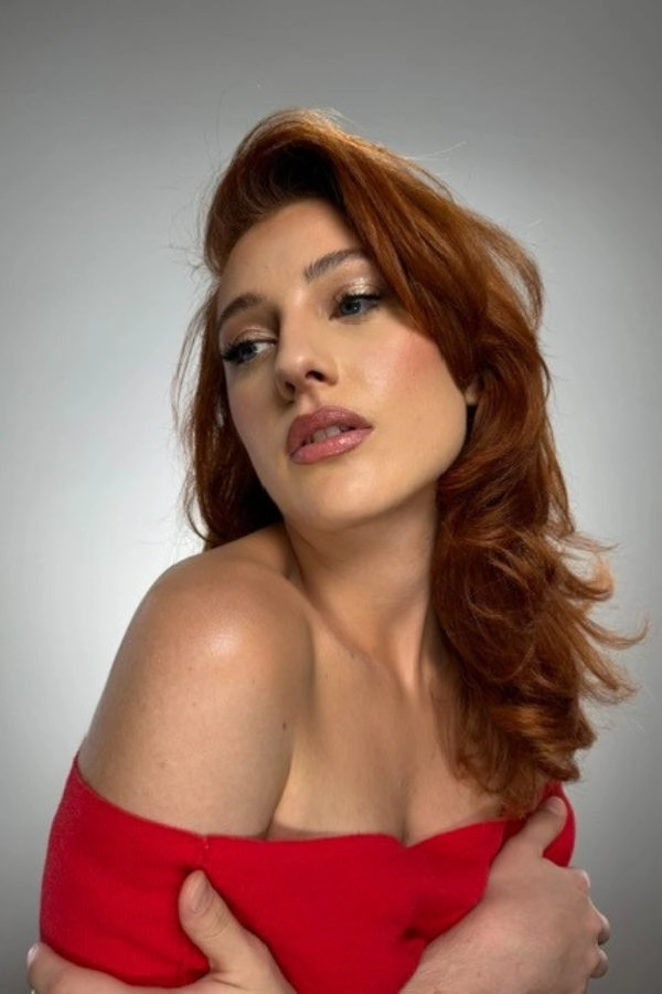 Γυναικείο μακιγιάζ με λαμπερή βάση, φυσικούς τόνους στα χείλη και απαλή σκιά ματιών, συνδυασμένο με κυματιστά κόκκινα μαλλιά για εντυπωσιακό και θηλυκό look.