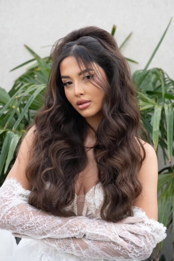 Νυφικό χτένισμα half up με πλούσιους κυματισμούς, ανάλαφρο όγκο και curtain bangs, συνδυασμένο με ρομαντικό νυφικό δαντέλας για θηλυκή bridal εμφάνιση.