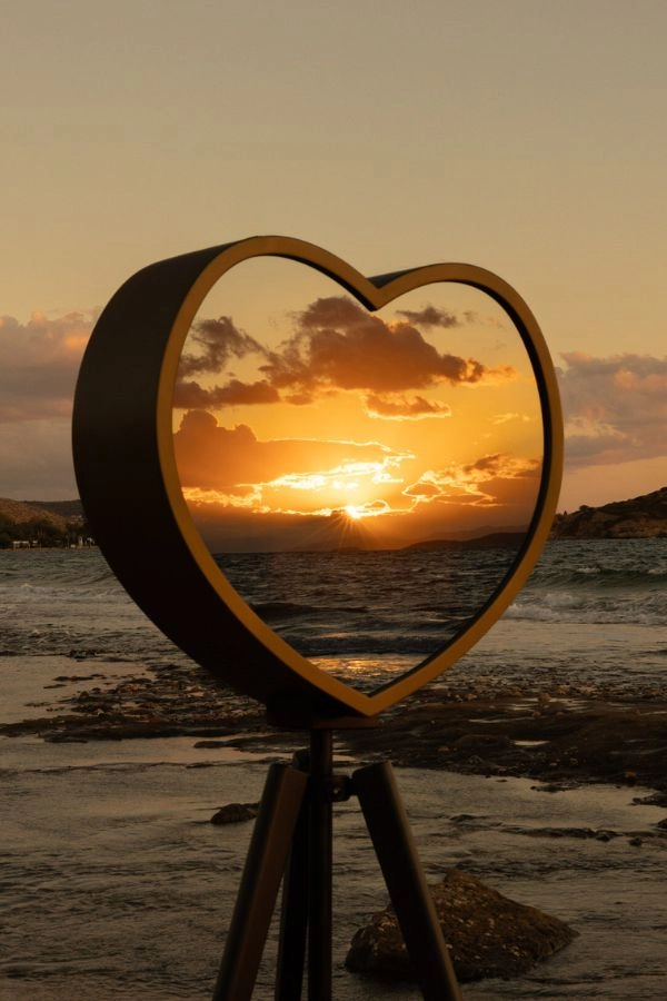 Mirror booth σε σχήμα καρδιάς με φόντο ηλιοβασίλεμα στη θάλασσα