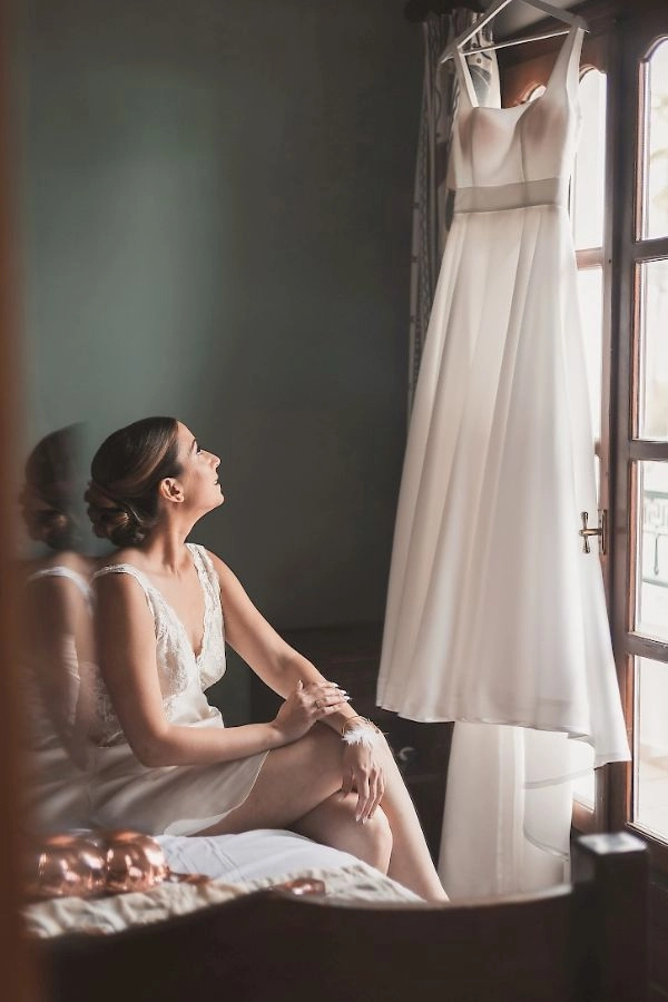 Νύφη στην προετοιμασία του γάμου κοιτά το νυφικό της δίπλα στο παράθυρο
