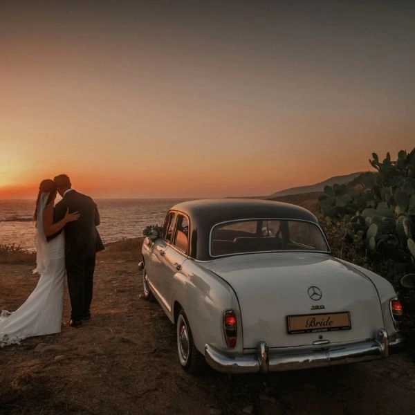 Νεόνυμφο ζευγάρι δίπλα σε κλασικό αυτοκίνητο γάμου Mercedes σε ρομαντικό ηλιοβασίλεμα