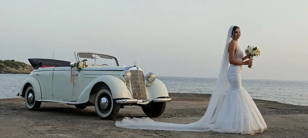 Νύφη με μακρύ πέπλο δίπλα σε κλασικό αυτοκίνητο γάμου σε παραθαλάσσιο τοπίο