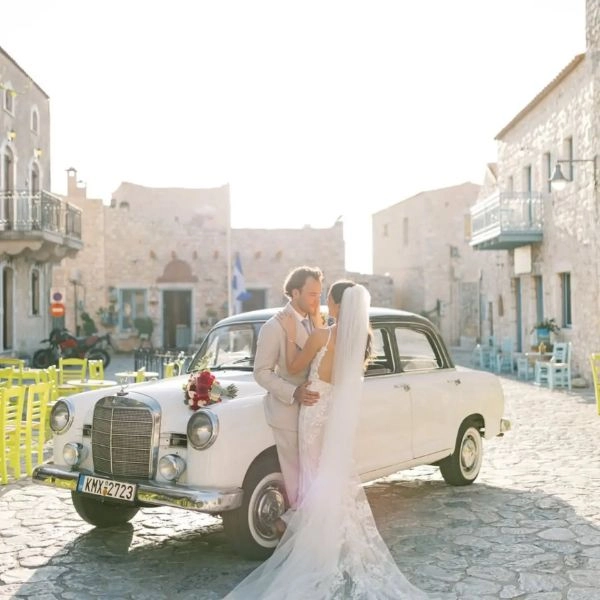 Νύφη και γαμπρός μπροστά από κλασικό αυτοκίνητο γάμου σε παραδοσιακό πέτρινο χωριό